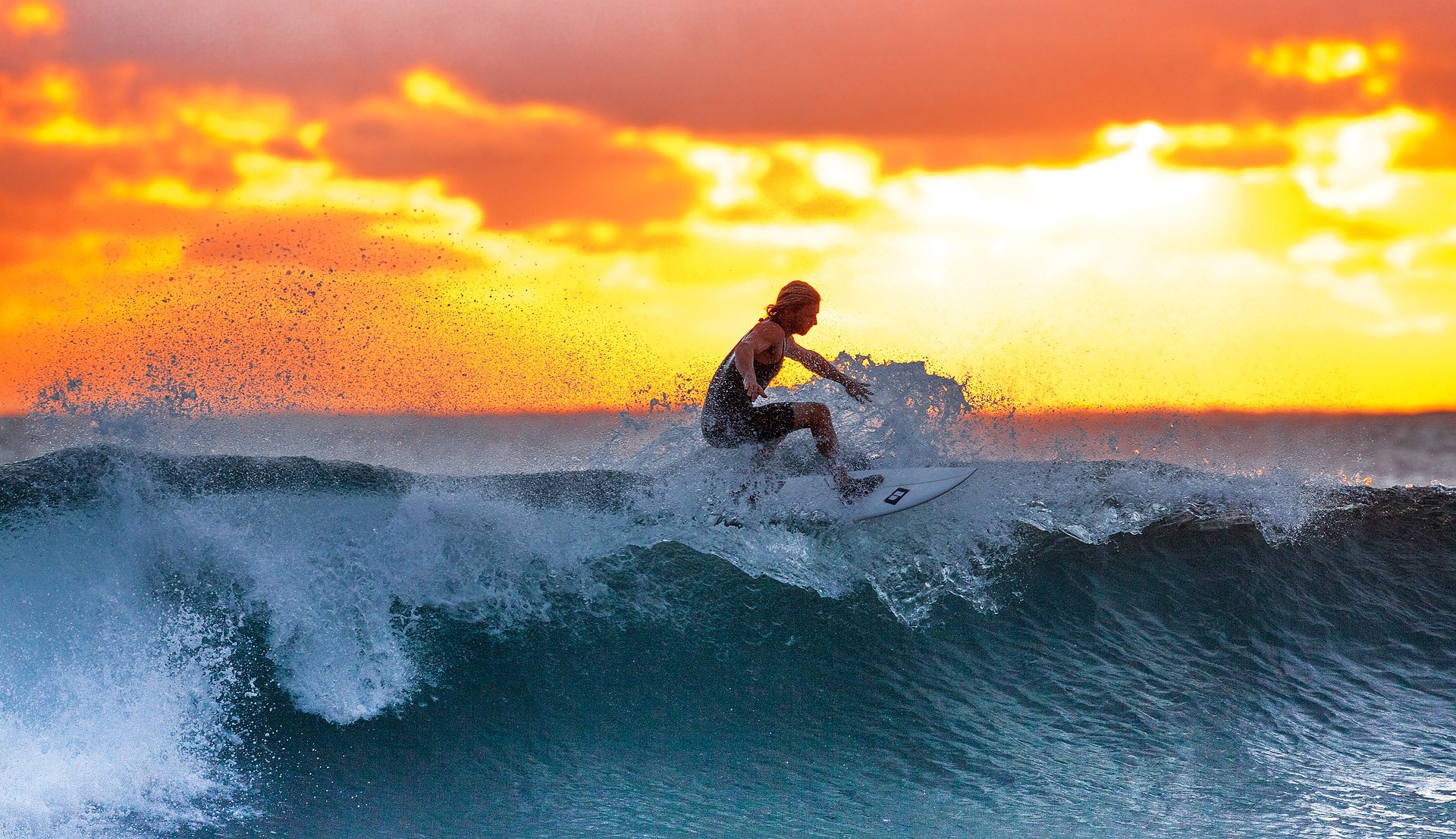 A surfer stands strong on their surfboard, riding the wave with a sunset glowing behind him. 